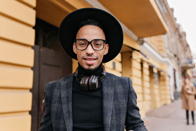 散步疲惫的非洲男模戴着大耳机站在餐厅附近的特写照片黑皮肤的自信帅哥的户外照片帽子寒冷成人