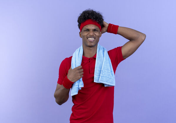 印象令人印象深刻的年轻美国黑人运动男子戴头带和腕带把手放在头上与毛巾在肩上隔离在蓝色的墙壁墙腕带肩膀