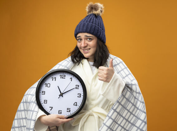 表演带着微笑的生病的年轻女子 戴着裹着格子布的长袍 戴着冬天的帽子 拿着钟 看着前面 竖起大拇指 孤零零地站在橙色的墙上人抱着钟