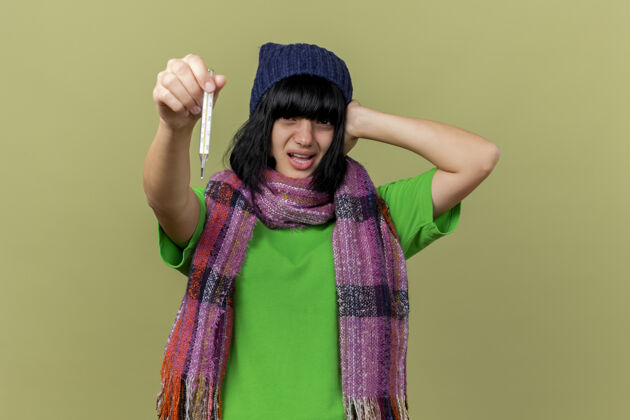 围巾戴着冬日帽 戴着围巾 焦急的年轻病妇把体温计伸到前面 手放在头上 望着橄榄绿墙上孤立的前方朝向站立手