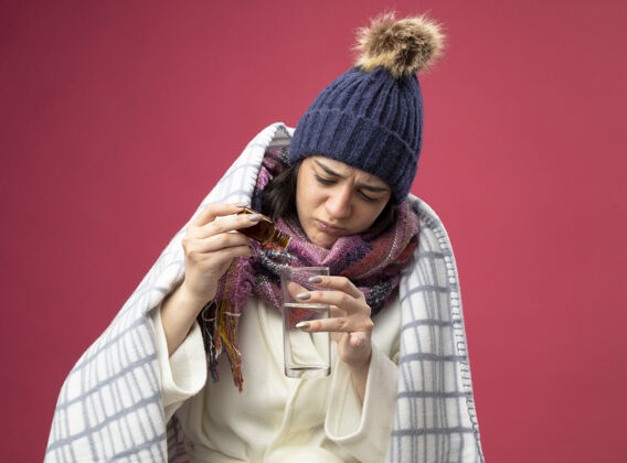 衣服专注的年轻病妇穿着长袍 戴着冬天的帽子 围巾裹着格子布 往隔离在粉红色墙上的一杯水里加药人注意力疾病
