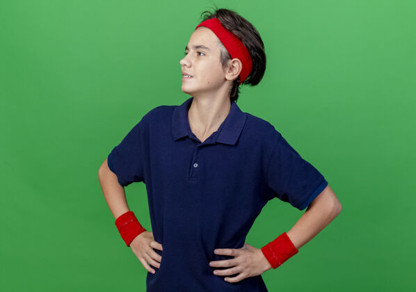 感情自信的年轻帅气的运动男孩戴着头带和护腕 戴着牙套 手放在腰上 看着绿色墙壁上孤立的一面头带腰穿