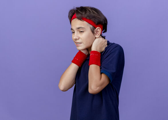 脸年轻帅气的运动男孩 戴着头带和护腕 戴着牙套 看起来笔直 手放在脖子后面 隔离在紫色的墙上脖子站人