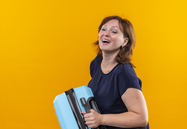 手提箱看着身边快乐的中年旅人女人拿着手提箱孤零零地站在黄色的墙上中年欢乐壁板