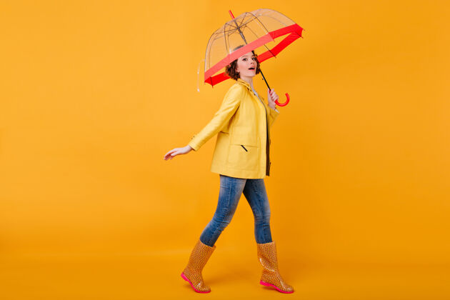 年轻穿着黄色夹克和胶鞋的白人女模特的全长肖像无忧无虑的卷发女孩打着伞跳舞的摄影棚镜头阳伞女人微笑
