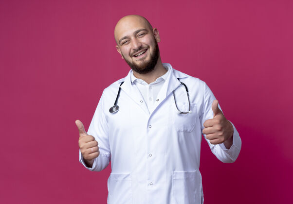 年轻微笑着的年轻男医生穿着医用长袍和听诊器 他的大拇指孤立地竖起在粉红色的皮肤上微笑穿拇指