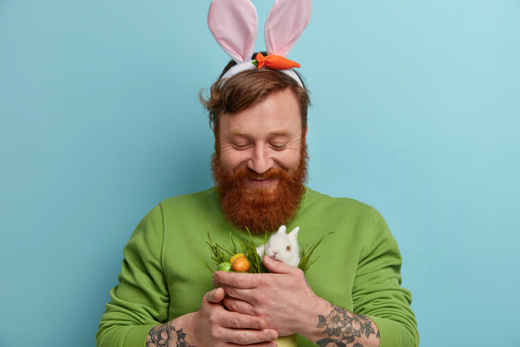 胡须复活节快乐留着胡子的姜人戴着兔子耳朵 抱着装饰着彩色彩蛋的小白兔 在春天庆祝宗教节日 对着蓝色的墙壁摆姿势寻蛋脸事件油漆