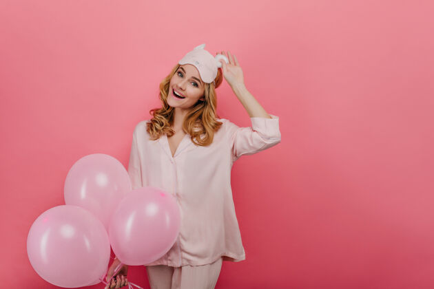 氦气球无忧无虑的白人妇女触摸着眼罩 在粉红色的墙上笑着穿着睡衣的漂亮生日女孩拿着气球享受着聚会微笑节日美丽
