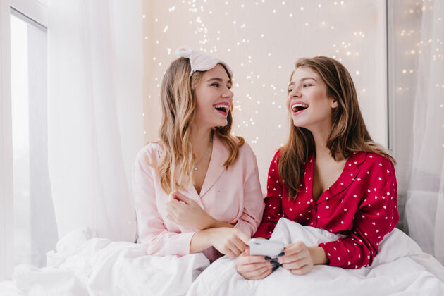 床穿着睡衣的女孩们面带微笑地看着对方高加索女士们坐在床上拿着智能手机笑的室内照片舒适年轻快乐