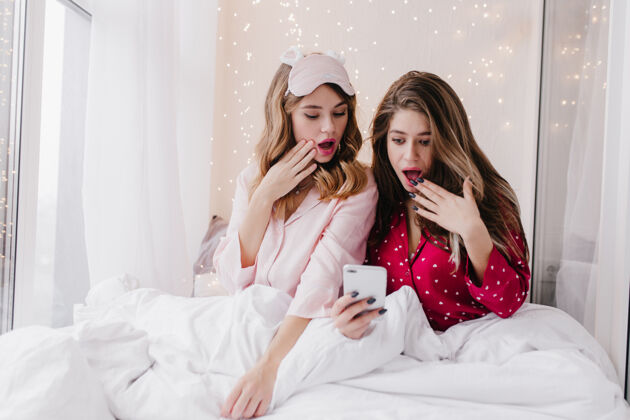 寒冷放松的女孩在床上拍照时表达惊讶的情绪令人惊讶的女性朋友用智能手机坐在白色毯子下的室内照片室内金发电话