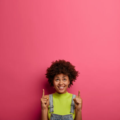 指示器看那里相当满意的女人有快乐的表情 促销商品 上面标明 在这里说你的促销 在粉红色的墙上展示你的广告内容的复制空间春季销售种族女性外表