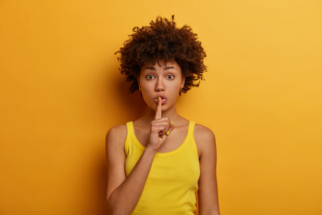 无语沉默和秘密的概念惊讶的非裔美国妇女把食指按在嘴唇上 要求安静 告诉非常私人的信息 穿着休闲的黄色背心 摆姿势 说嘘或嘘青年沉默穿着