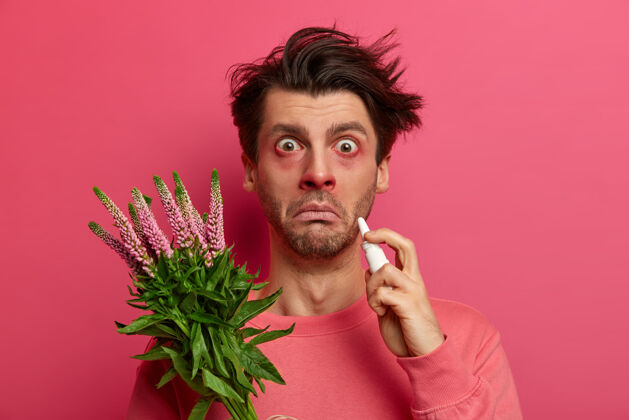 花粉患有过敏性疾病的年轻人滴鼻液 眼睛和鼻子发红 对植物过敏 鼻炎或花粉热症状 凝视 对着粉红色的墙壁摆姿势 对花粉有反应肿胀攻击过敏