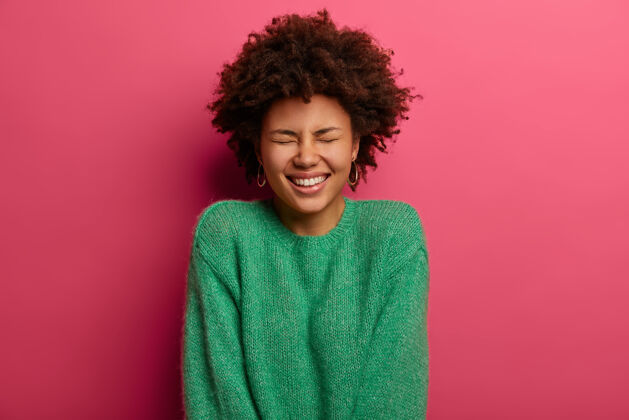 感觉快乐情绪化的卷发女孩咧嘴笑 表达积极的情绪 闭上眼睛不笑 穿着绿色的毛衣 感觉乐观 孤立在粉红色的墙上 散发着快乐真诚种族乐观
