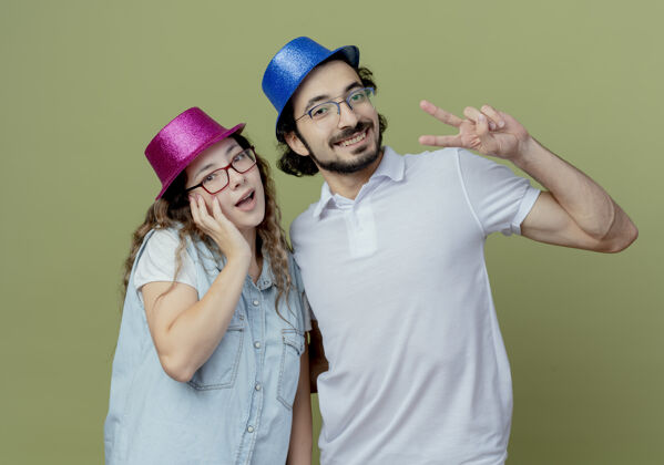 微笑微笑的年轻夫妇戴着粉色和蓝色的帽子女孩把手放在脸颊上 男孩表现出和平的姿态孤立在橄榄绿上年轻橄榄情侣