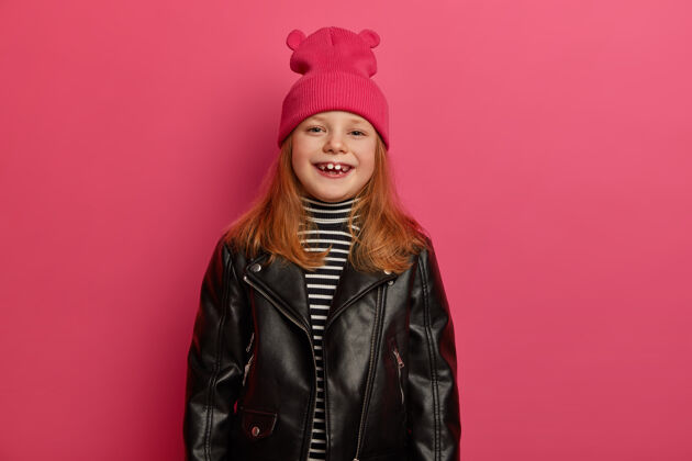 欧洲积极微笑的红头女孩积极地笑着 戴着粉红色的帽子和皮夹克 看起来很高兴 隔着粉红色的墙孩子们 时尚概念可爱的女儿喜欢购物搞笑小生姜