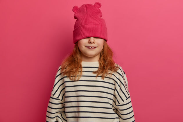 牙齿有趣的小姜女躲在时髦的帽子后面 露出两颗乳牙 穿着条纹休闲套头衫 在室内玩得很开心 孤立在粉色的墙上 是个淘气的孩子快乐童年的概念欧洲人乐观