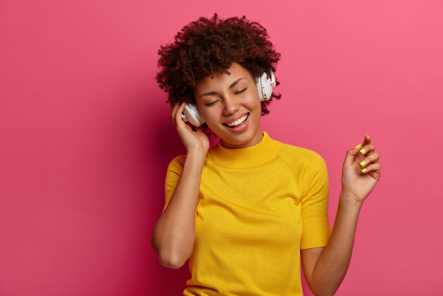 非洲乐观的少数民族女孩无忧无虑地移动 在耳机里听音乐 感觉轻松 在歌曲应用程序上享受最喜欢的旋律或新曲目 穿着黄色衣服 隔离在粉红色的墙上科技 小玩意兴奋快乐心情