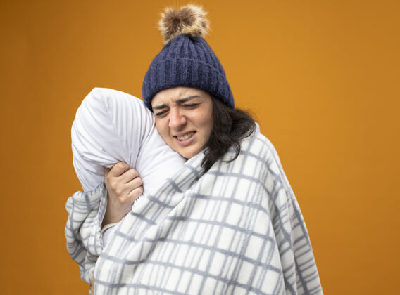 感觉疼痛的年轻生病的女人穿着长袍 戴着冬天用格子布包裹的帽子 站在侧视图里 抱着枕头 闭着眼睛孤立地站在橙色的墙上女人冬天人