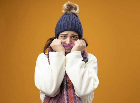 年轻人焦急的年轻生病的女人穿着长袍 戴着冬天的帽子和围巾 看着前面用围巾遮住的嘴巴隔离在橙色的墙上姿势疾病嘴