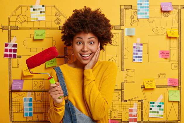 房间漂亮高兴的黑皮肤女人忙着房子的重新装修 搬进新公寓 拿着油漆辊 油漆完墙后休息 穿着黄色毛衣和工作服 站在设计草图上刷子女人家庭