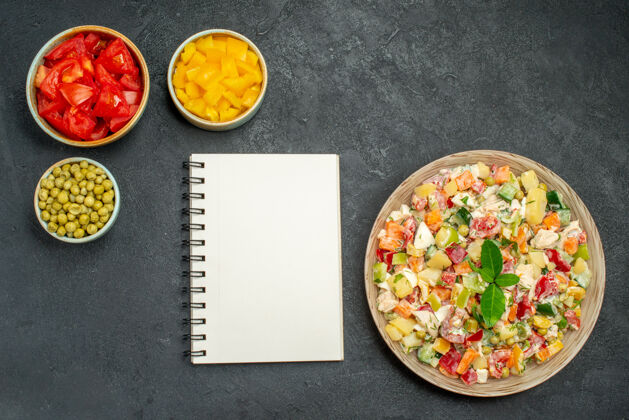 菜肴深灰色背景上蔬菜沙拉的顶视图 旁边有碗蔬菜和记事本一餐晚餐烹饪