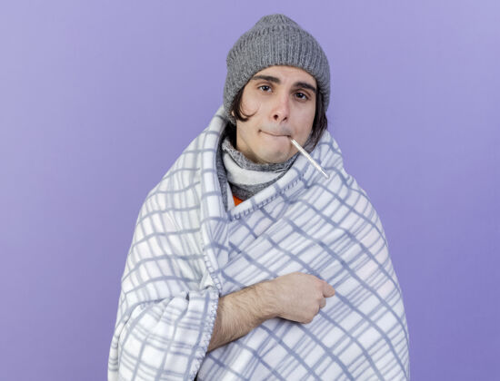 虚弱看着镜头虚弱的年轻病人戴着冬天的帽子 围巾裹着格子布 把体温计放进嘴里 隔离在紫色的背景上格子围巾温度计