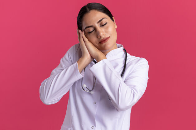 头年轻的女医生 穿着医用外套 手持听诊器 做着睡觉的姿势 双手并拢 头靠在手掌上 站在粉红色的墙上使站立年轻