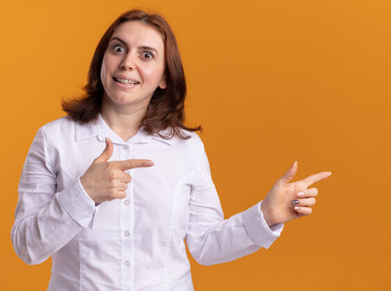 女人身穿白衬衫的年轻女子站在橘色的墙上 面带微笑 用食指指着一旁站立微笑手指