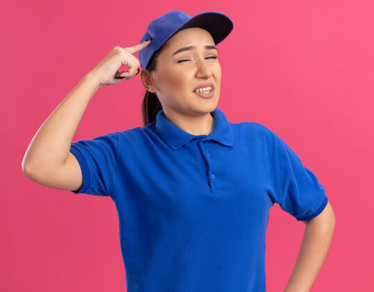 制服穿着蓝色制服 戴着帽子的年轻女送货员站在粉红色的墙上抓着头 看上去很困惑站立头女人