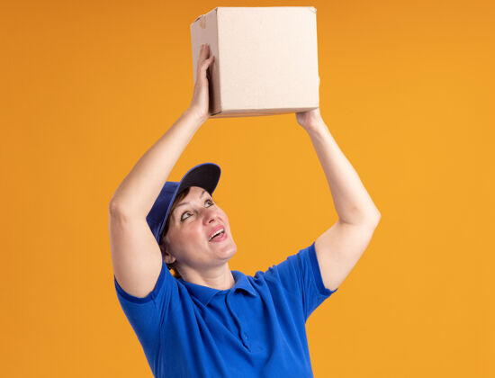 制服身穿蓝色制服 头戴鸭舌帽的中年送货妇女站在橙色的墙上 头上顶着一个纸板箱 面带微笑地看着它年龄盒子中间