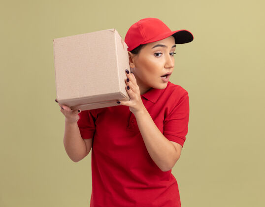 拿着年轻的送货员身穿红色制服 头戴鸭舌帽 耳朵上放着纸板箱 站在绿色的墙上听着女人帽子站着