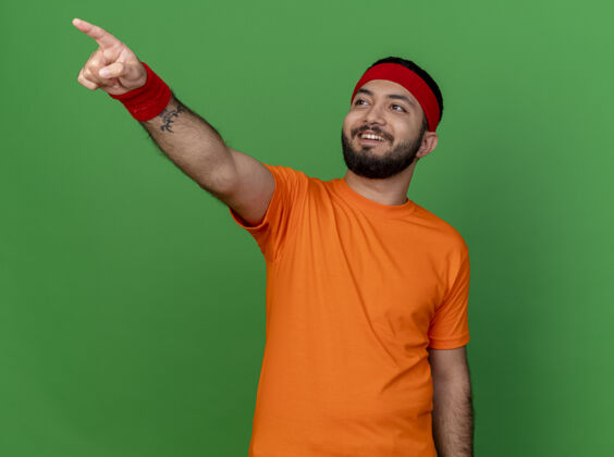 运动面带微笑的年轻运动型男子看着戴着头带和腕带的一侧 手指孤立在绿色背景上尖年轻壁板