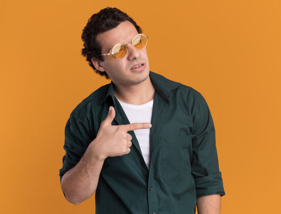 年轻人一个身穿绿衬衫 戴着眼镜的年轻人面带严肃的表情 用食指指着站在橙色墙上的一边目录站靠边