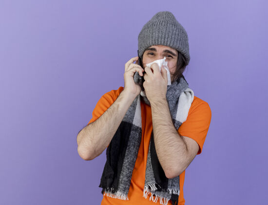 鼻子戴着带围巾的冬帽 用紫色背景隔离的餐巾纸擦鼻子的年轻病患在电话里讲话餐巾帽子电话