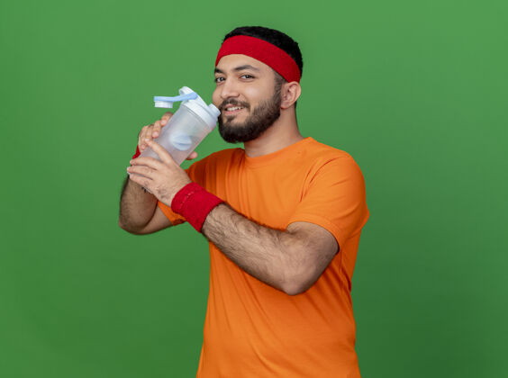 腕带戴着头带和腕带的年轻运动型男子微笑着从绿色背景上孤立的水瓶里喝水微笑年轻运动
