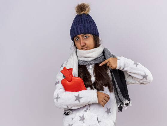包年轻的生病的女孩看着相机戴着冬天的帽子 拿着围巾 指着孤立在白色背景上的热水袋女孩点围巾