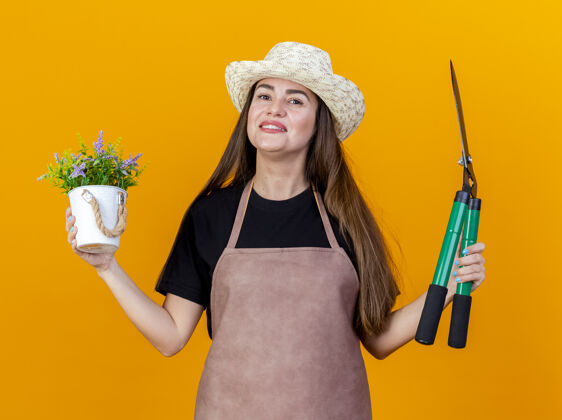 花微笑美丽的园丁女孩穿着制服 戴着园艺帽 拿着剪子在花盆里拿着花 摊开的手孤立在橙色的背景上摊开花盆手捧