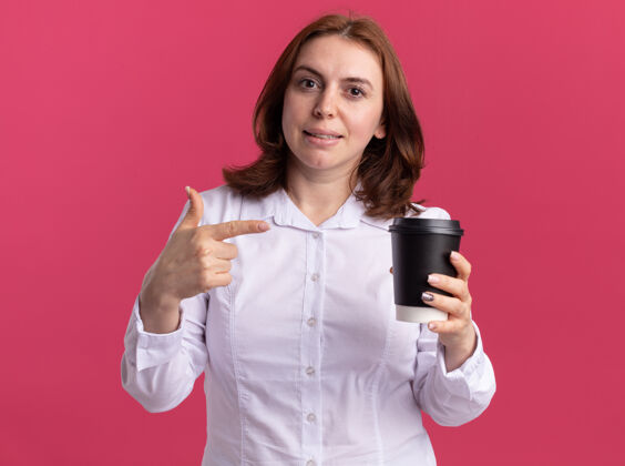 女人身穿白衬衫的年轻女子拿着咖啡杯 食指指着咖啡杯 自信地微笑着站在粉红色的墙上拿着杯子咖啡