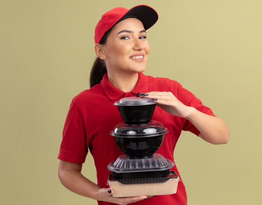 女人身着红色制服 戴着帽子的快乐的年轻女送货员 手里拿着一叠食品包 面带微笑地站在绿色的墙边看着前方制服递送帽子