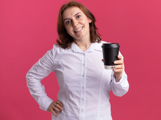 年轻穿着白衬衫的快乐的年轻女子拿着咖啡杯看着前面 站在粉红色的墙上 开心地微笑着站着衬衫杯子