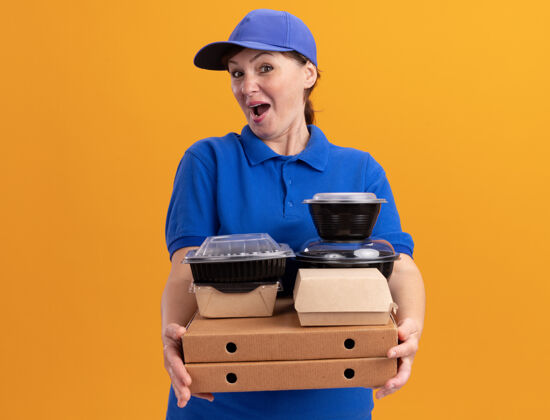 抱着快乐的中年送货员身穿蓝色制服 戴着帽子 手里拿着披萨盒和食品包 站在橘色的墙上 面带微笑地看着前面女人制服中年