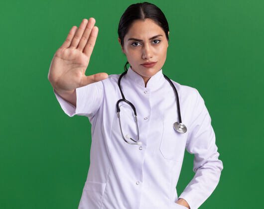 医生年轻的女医生 穿着医用外套 脖子上戴着听诊器 严肃地看着前方 张开的手站在绿色的墙上 做着停止的手势外套脸严重