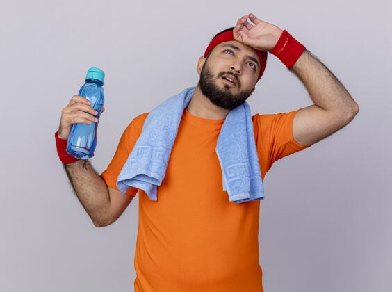 年轻疲惫的年轻运动型男子抬头戴着头带和腕带 肩上拿着毛巾的水瓶 手放在额头上 隔离在白色背景上累了手瓶子