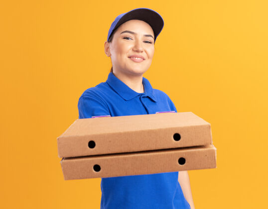 站着快乐的年轻送货员身穿蓝色制服 戴着帽子 手里拿着比萨饼盒 微笑着自信地站在橙色的墙上看着前面盒子自信年轻