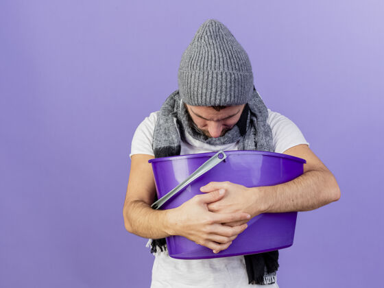 疾病戴着带围巾的冬帽的年轻病夫低着头 手里拿着塑料筐 把呕吐物放在紫色的背景上隔离开来篮子降低帽子