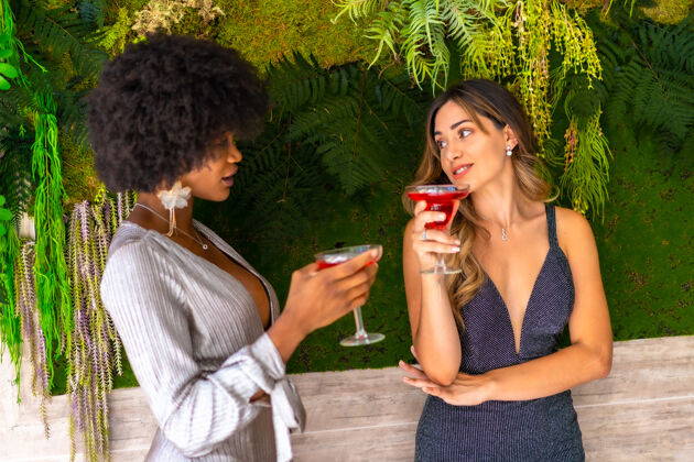 饮酒非裔美国人和白人女性朋友穿着花哨的衣服喝酒聊天多民族鸡尾酒享受
