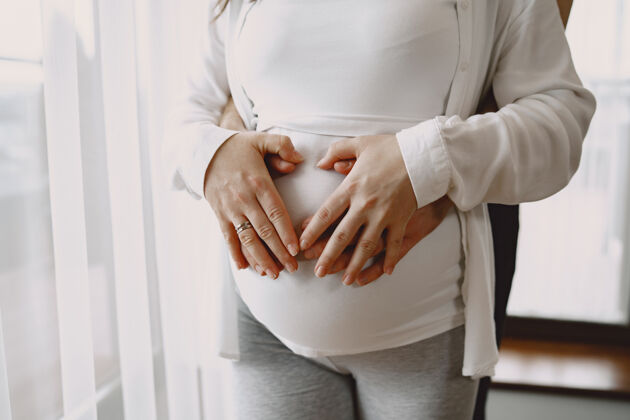 男人穿着轻便衣服的孕妇手放在孕妇的肚子上靠窗的家人支持怀孕金发