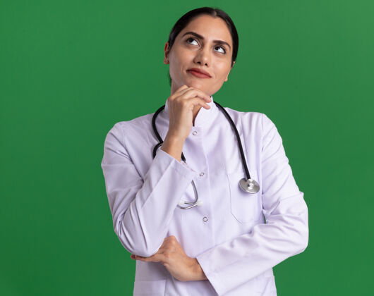 听诊器身穿医用外套 脖子上戴着听诊器的年轻女医生抬起头沉思着站在绿色的墙上医学脖子站