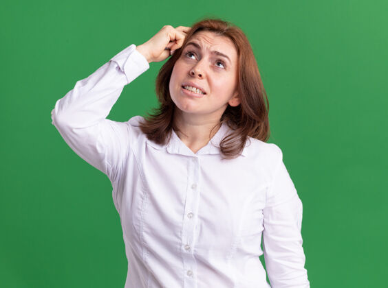 头穿白衬衫的年轻女子站在绿色的墙上 困惑地抬起头来抓头年轻女人抓挠